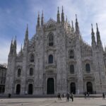 File in una strada affollata di Milano con traffico e persone in movimento.