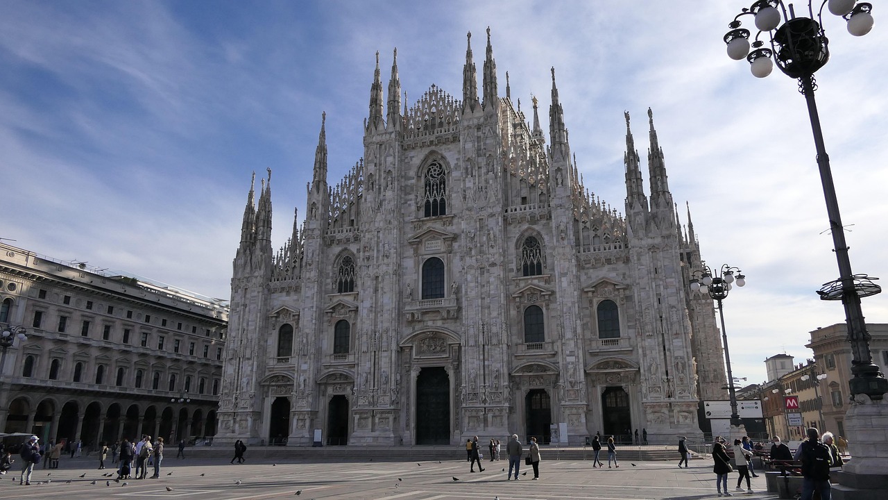 File in una strada affollata di Milano con traffico e persone in movimento.